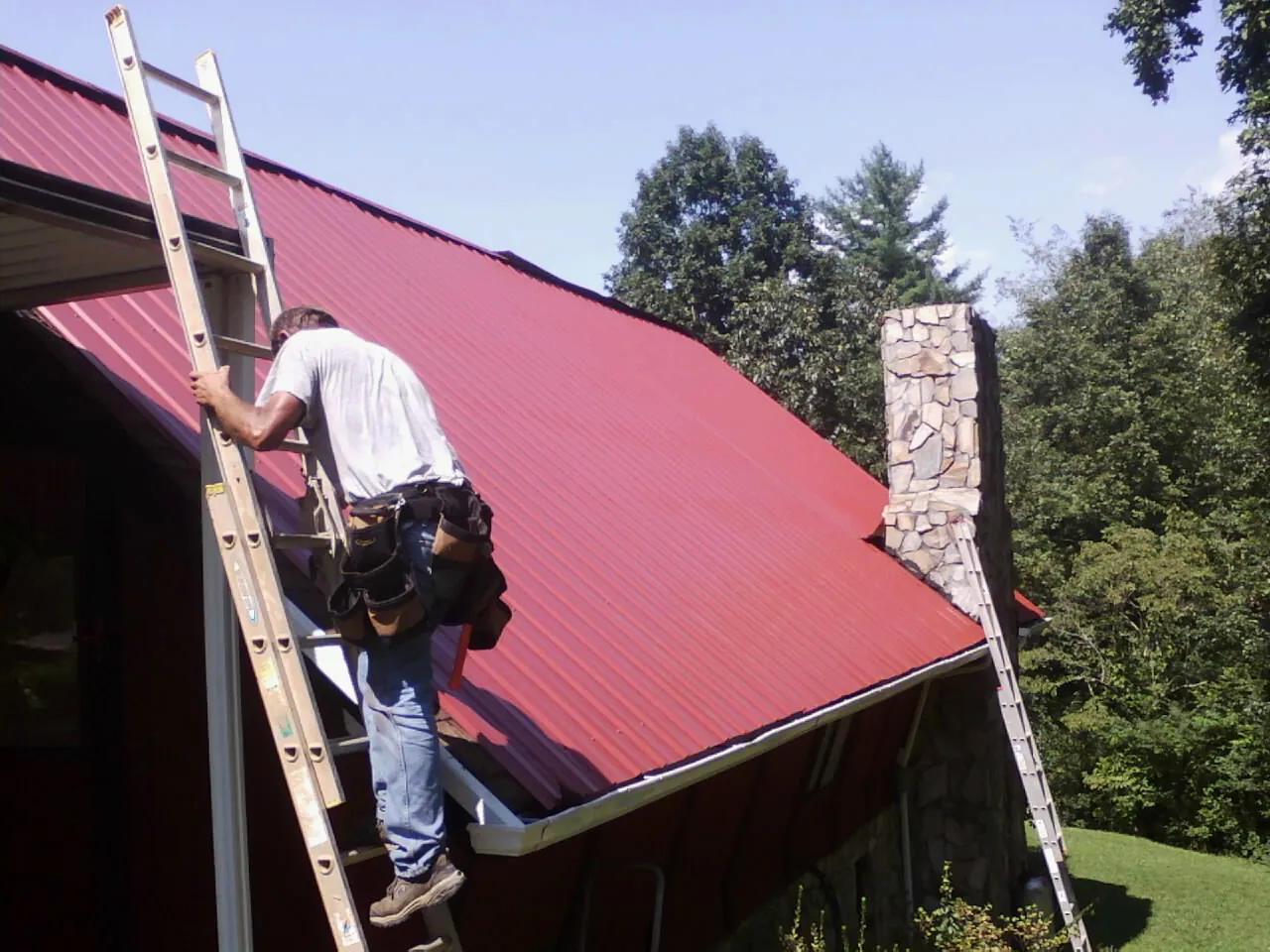 Roofer on a ladder beside a red metal roof and stone chimney — Soffit & Fascia Repair by Roofing Brothers in Deep Run